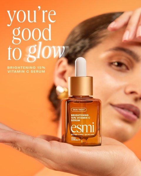 A close-up portrait of a model holding a Vitamin C serum bottle against a warm orange backdrop, focusing on a healthy, radiant skin aesthetic.